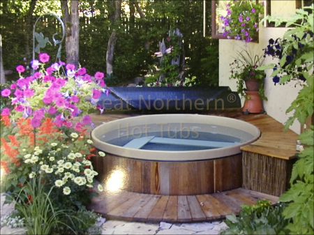 <p>Great Northern Rubadub Tub western red cedar hot tub with a rolled-up cover nearby with small surrounding deck in a garden setting.</p>