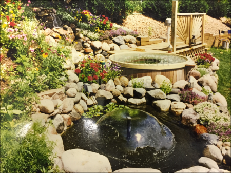 <p>Outdoor round wooden hot tub with small deck in a rock garden and pond.</p>
